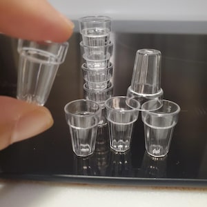 May include: Several small, clear plastic shot glasses are arranged on a dark surface, with one held by a finger. The glasses are simple in design, with straight sides. They are suitable for miniature drinks or decorative use.