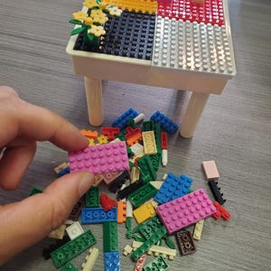 May include: A small white plastic table with a building block surface featuring yellow, red, black, and white sections. A small yellow flower arrangement sits on the black section. A pile of assorted colorful plastic building bricks lies below.