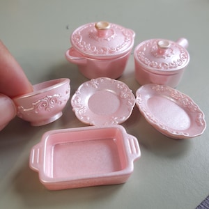 May include: A set of miniature kitchenware in a pearlescent light pink finish, including two lidded pots, a small bowl, a round plate, an oval platter, and a rectangular baking dish, all featuring ornate embossed scrollwork along the edges.