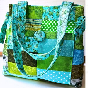 May include: A patchwork tote bag constructed from various fabric squares in shades of green and blue. The design features patterns of frogs, dragonflies, and white polka dots. A fabric-covered button sits at the center, and two matching shoulder straps are attached to the top.