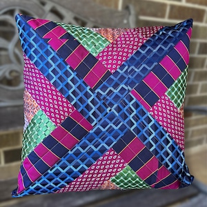 May include: A square throw pillow constructed from a patchwork of silk neckties. The design features a woven, crisscross pattern of strips in vibrant shades of magenta, royal blue, and emerald green, accented with various geometric and polka dot prints.
