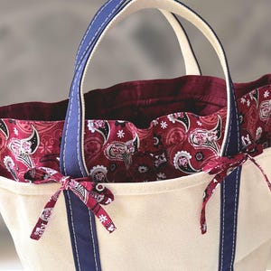 May include: A cream-colored canvas tote bag featuring dark blue fabric handles and trim. The interior is lined with a deep red fabric decorated with a white and black paisley pattern. Two small fabric ties in the same paisley print are attached.