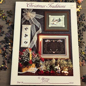 May include: A Stoney Creek Collection Book 28 titled Christmas Traditions featuring cross-stitch patterns. The cover displays framed embroidery, a vertical banner with bird motifs, a basket of red roses, and lace accents on a dark background.