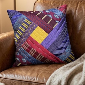 May include: A square throw pillow constructed from a patchwork of various neckties in shades of purple, burgundy, yellow, and blue. The fabric strips are arranged in a diagonal pattern, featuring diverse textures and geometric prints.