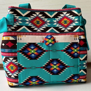 May include: A tote bag featuring a vibrant turquoise, red, yellow, and black geometric pattern. The bag includes two turquoise side ties, a front pocket secured with a matching fabric-covered button, and two turquoise shoulder straps.
