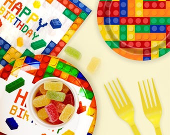 May include: A festive birthday party set featuring plates, napkins, and forks with a colorful block design. The plates have a white center with the words "Happy Birthday". The set includes a small bowl of colorful gummy candies.