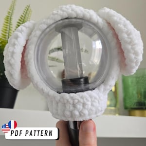 May include: A white, fluffy, crocheted bunny ear cover for a light stick. The cover is made of soft, textured yarn and features pink inner ears. The light stick is clear with a black base. The image includes the text "PDF PATTERN".