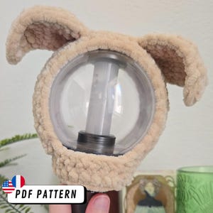 May include: A beige, plush, bunny ear cover for a clear, spherical light. The cover is made of soft, textured material and has two upright ears. The light has a cylindrical center. The text "PDF PATTERN" is visible.