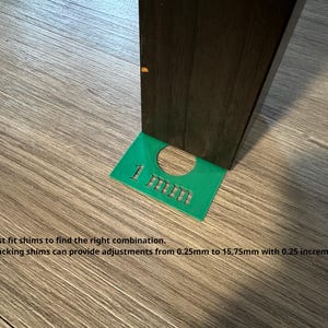 May include: A green rectangular shim with a circular cutout and the text 1 mm embossed on the surface. The shim is positioned under the dark brown leg of a piece of furniture. Text on the image reads: Test fit shims to find the right combination. Stacking shims can provide adjustments from 0.25mm to 15.75mm with 0.25 increments.