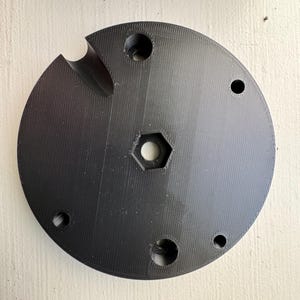 Puede incluir: Un componente negro y circular con un orificio central hexagonal y varios orificios circulares más pequeños. Una muesca curva está cortada en el borde. La superficie tiene un patrón texturizado y estriado.