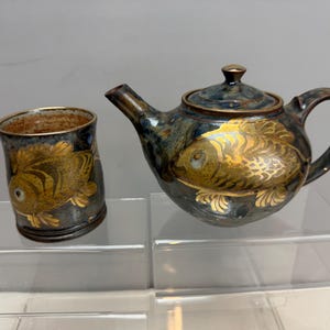 May include: A ceramic teapot and matching cup set featuring a dark blue and grey mottled glaze with metallic gold koi fish illustrations. The teapot has a curved spout, a lid with a small knob, and a handle, while the cup is cylindrical.