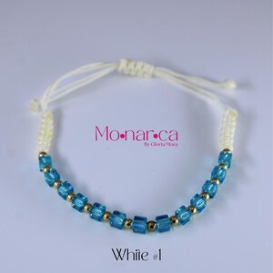 May include: A white cord bracelet with blue square beads and gold-colored accents. The bracelet has the text "Mo.nar.ca" and "White #1". The bracelet is adjustable.