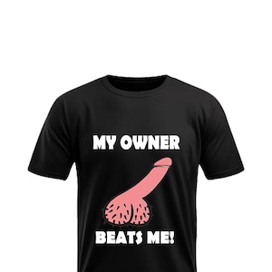 以下が含まれることがあります： 黒いTシャツ。前面中央にピンク色のペニスのグラフィックと白い文字で「MY OWNER BEATS ME！」と書かれています。デザインはTシャツの中央に配置されています。