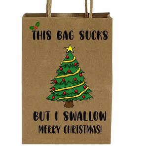 Könnte beinhalten: Eine braune Papiertüte mit gedrehten Papiergriffen. Die Tüte zeigt eine Weihnachtsgrafik und den Text "THIS BAG SUCKS BUT I SWALLOW MERRY CHRISTMAS!" in Schwarz. Ein Stechpalmenzweig befindet sich in der oberen linken Ecke.