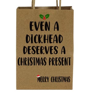 May include: A brown paper gift bag with twisted paper handles. The bag features the text "EVEN A DICKHEAD DESERVES A CHRISTMAS PRESENT" in black, with a sprig of holly and the words "MERRY CHRISTMAS".