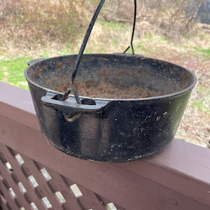 Könnte beinhalten: Ein schwarzer gusseiserner Dutch Oven mit einem gebogenen Metallgriff. Der Topf steht auf einem braunen Holzgeländer mit einem natürlichen Hintergrund aus Bäumen und Gras.