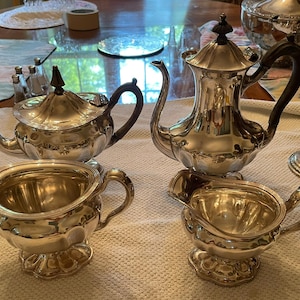 Op de afbeelding: Een zilveren theeservies met een theepot, melkkan, suikerpot en koffiepot. De theepot en koffiepot hebben zwarte handvatten. De set staat op een wit tafelkleed.