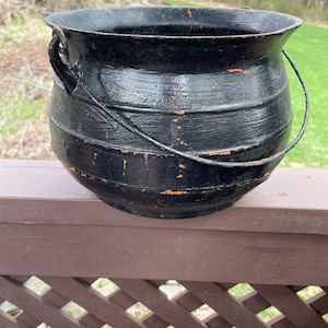 May include: A black cast iron cauldron with a handle and a wire bail. The cauldron has a rounded bottom and horizontal bands around the body. The paint is chipped, revealing orange underneath. A rustic, vintage piece.