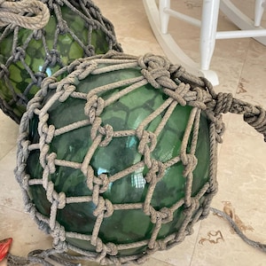May include: Two green glass fishing floats encased in knotted rope netting. The floats are round and have a vintage, nautical aesthetic. The rope is a neutral beige color, and the glass has a slight green tint. The floats are on a tiled surface.