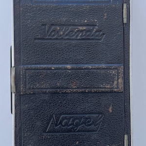 May include: A vintage black leather-covered folding camera with a rectangular body and a metal frame. The front features the embossed text Vollenda and Nagel, along with a small circular red window. A worn leather carrying strap is attached to the top.