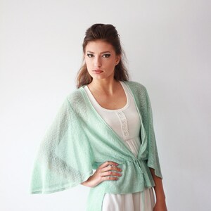 May include: A light green open-front cardigan with a tie at the waist. The cardigan is made of a lightweight, airy fabric and has a relaxed fit. The model is wearing a white tank top underneath the cardigan.