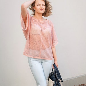 May include: A light pink, open-weave top with short sleeves and a round neckline. The top is paired with light blue jeans and a black, navy, and beige handbag. The woman is holding her hair up with one hand.