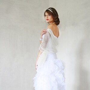 May include: A white bridal shawl with a delicate lace pattern, worn over a white dress with a ruffled skirt.