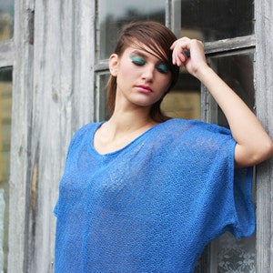 May include: A woman wearing a blue, loose-fitting, short-sleeved top. The top has a scoop neck and a textured, open-knit design. The background features a weathered wooden wall and window.