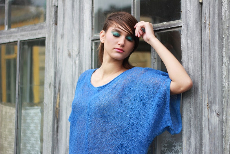 May include: A woman wearing a blue, loose-fitting, short-sleeved top. The top has a scoop neck and a textured, open-knit design. The background features a weathered wooden wall and window.
