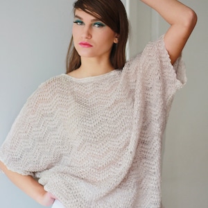 May include: A woman wearing a loose-fitting, beige knitted sweater with a delicate open weave. The sweater has a round neckline and short sleeves. The woman is wearing a white skirt and has her hair pulled back.