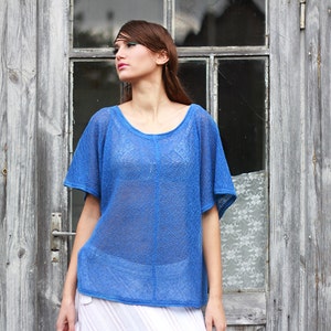 May include: A blue, short-sleeved top with a scoop neck. The top is made of a sheer, open-weave fabric. The model is wearing a white skirt. The background is a weathered wooden door.
