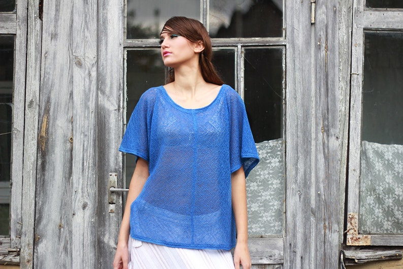 May include: A blue, short-sleeved top with a scoop neck. The top is made of a sheer, open-weave fabric. The model is wearing a white skirt. The background is a weathered wooden door.