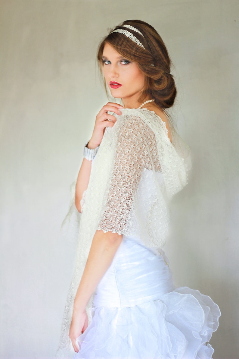 May include: A white lace shawl with a delicate pattern. The shawl is draped over a white dress with a ruffled skirt. The model is wearing a pearl necklace and a headband with a white lace design.