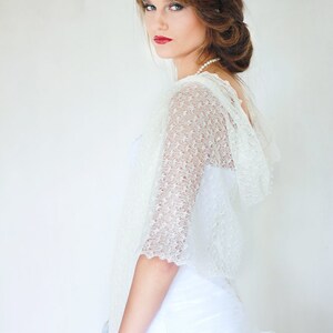 May include: A white lace bridal shawl with a delicate pattern. The shawl has a hood and is worn over a white dress.