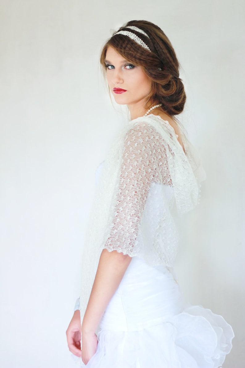 May include: A white lace bridal shawl with a delicate pattern. The shawl has a hood and is worn over a white dress.