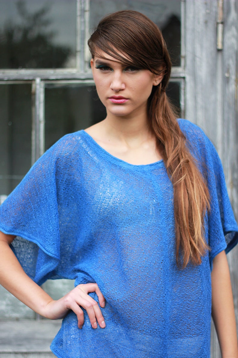 May include: A blue, short-sleeved top with a scoop neck. The top has a loose, flowing fit and is made of a sheer, textured fabric. The model is wearing the top with her hand on her hip.