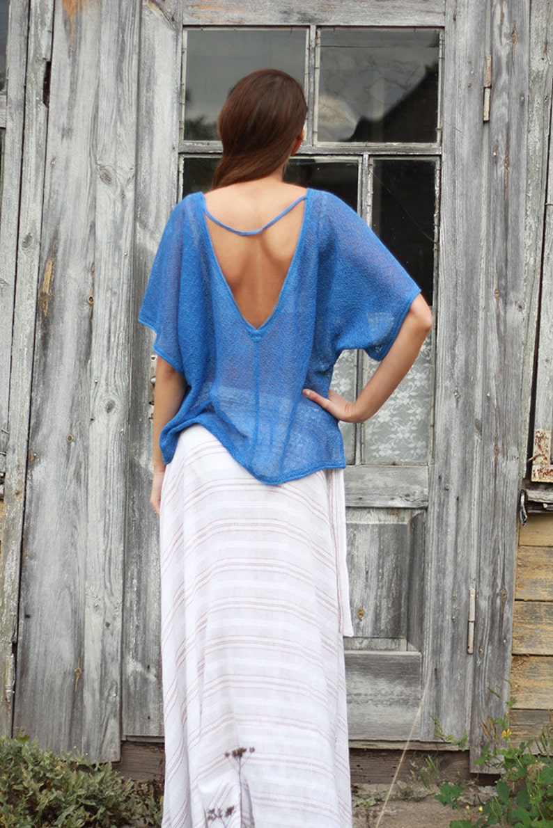 May include: A blue open-back top with short sleeves and a deep V-neck. The top is paired with a long, white and light brown striped skirt. The clothing is worn in front of a weathered wooden door.