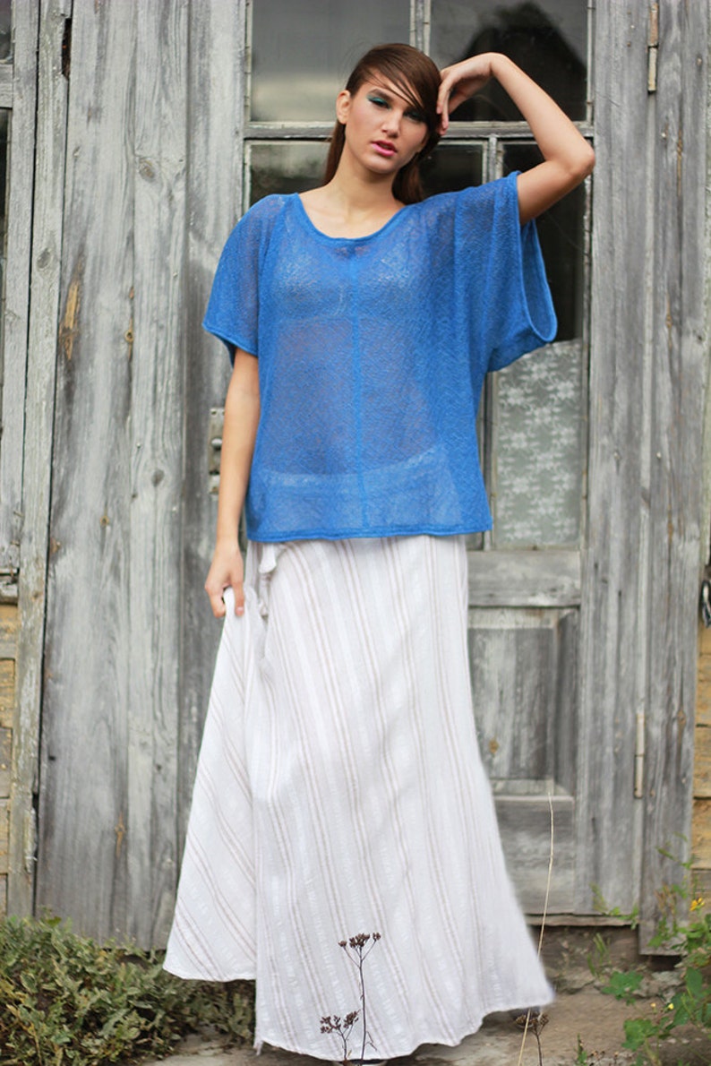 May include: A woman's outfit featuring a sheer, short-sleeved, royal blue top and a long, flowing white skirt with vertical stripes. The clothing is lightweight and appears suitable for warm weather. The background is a weathered wooden door.