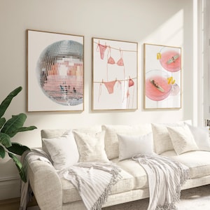 May include: Three framed art prints. The first shows a disco ball, the second a pink bikini on a washing line, and the third two pink cocktails. The prints are above a cream sofa.