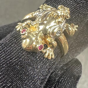 May include: A gold-toned ring in the shape of a frog, featuring red and clear gemstones. The frog's body is detailed with textured surfaces and small embellishments. The ring is shown on a dark background.