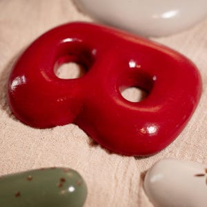 May include: A vibrant red, abstract ceramic piece with two holes, alongside other smooth, colourful ceramic shapes. The objects are displayed on a textured, beige fabric, forming a creative, artistic arrangement.