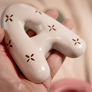 May include: A white ceramic letter "A" with a glossy finish and small, cross-shaped cutouts. The decorative piece is held in a hand, showcasing its smooth texture and unique design. The letter is a decorative item.