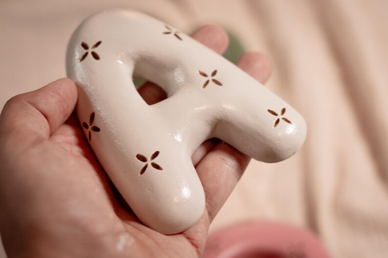 May include: A white ceramic letter "A" with a glossy finish and small, cross-shaped cutouts. The decorative piece is held in a hand, showcasing its smooth texture and unique design. The letter is a decorative item.