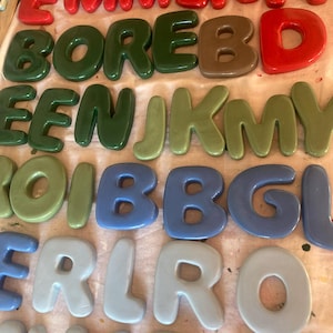 May include: A collection of colorful, glossy, 3D letters in various shades of red, green, brown, blue, and gray. The letters are arranged in rows, spelling out names and words. The letters are likely for decorative purposes.