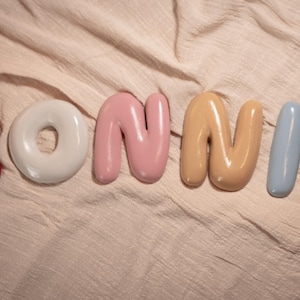 May include: A collection of colorful, glossy ceramic letters spelling "BONNIE". The letters are in various colors: red, white, pink, tan, light blue, and green. The letters are arranged on a textured, light-colored fabric.