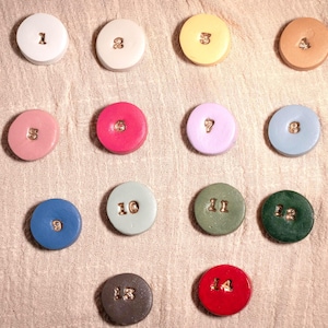 May include: A collection of fourteen colorful, round ceramic pieces, each with a different number from 1 to 14. The numbers are in a gold color. The pieces are arranged on a textured, light-colored surface.