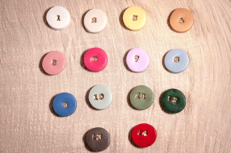 May include: A collection of fourteen colorful, round ceramic pieces, each with a different number from 1 to 14. The numbers are in a gold color. The pieces are arranged on a textured, light-colored surface.