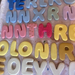 May include: A collection of colorful, bubble-style letters in various shades, including blue, pink, yellow, and green. The letters are arranged in rows, with some spelling out words. The letters have a glossy finish and are likely made of a hard material.
