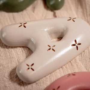 May include: A light-colored, ceramic decorative object in the shape of the number seven, with a hole in the middle. The object has a smooth surface and is decorated with small, cross-shaped cutouts. Other similar objects are visible in the background.