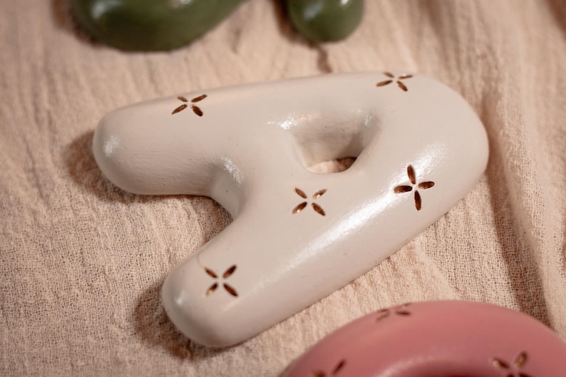 May include: A light-colored, ceramic decorative object in the shape of the number seven, with a hole in the middle. The object has a smooth surface and is decorated with small, cross-shaped cutouts. Other similar objects are visible in the background.